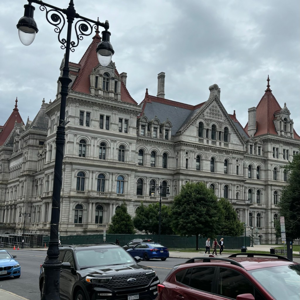 Historic building with ornate architecture featuring red rooftops and pointed turrets, seen from a street with parked cars in the foreground and a vintage lamppost. Cloudy sky in the background.