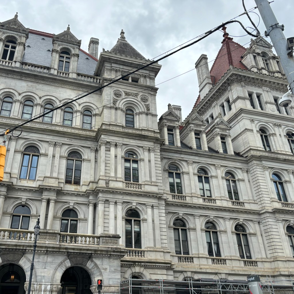 Historic building with ornate architectural details, grey stone facade, arched windows, and multiple peaked roofs. Cloudy sky in the background.