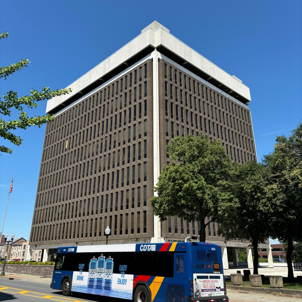 A tall, rectangular office building with a modern design stands against a clear blue sky. In the foreground, a blue city bus with advertisements for a beverage and an anti-drug message is driving past. Trees and a flagpole are also visible.